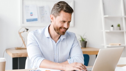 Person mit Bart und hellblauem Hemd sitzt freudig an einem Laptop während daneben ein aufgeschlagenes Notizbuch mit einem Stift und ein Smartphone liegt, im Hintergrund steht ein weiterer Schreibtisch, ein Regal sowie hängt eine Pinnwand an der Wand