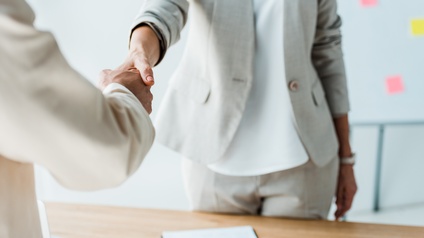 Zwei Personen in hellen Sakkos stehen sich an einem Tisch gegenüber und schütteln sich die Hände. Auf dem Tisch liegt ein Stift auf einem Zettel