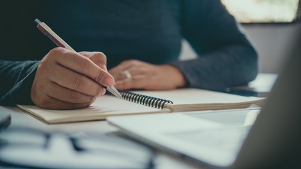 Eine Person sitzt an einem Tisch und hält einen Stift in der rechten Hand. Vor der Person liegt ein Notizbuch. Daneben ist ein aufgeklappter Laptop