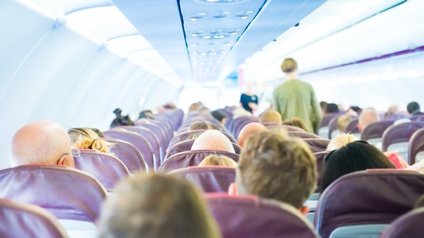 Innenraum eines Passagierfliegers in Rückenansicht: Mehrere Sitzreihen, auf denen Menschen sitzen, eine Person geht Gang entlang