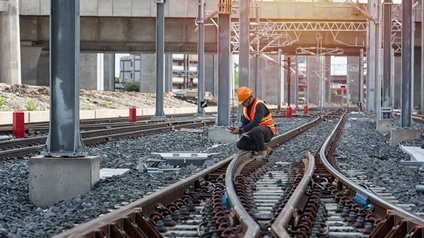 Eine Person mit Helm und Sicherheitsweste hockt auf Gleisen. Daneben sind weitere Gleise. Die Person hält ein Walkie Talkie
