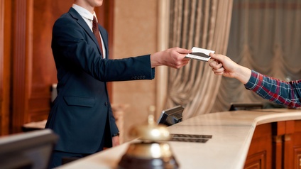 Person in Businesskleidung steht hinter einer noblen Rezeption mit dunklem rötlichen Holz und Marmorplatte und übergibt einer anderen Person eine Zimmerkarte