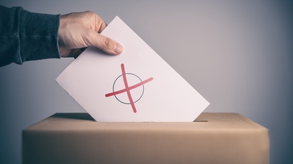Seitliche Aufnahme einer Wahlurne, in die eine Person einen weißen Zettel wirft, auf dem ein schwarzer Kreis ist, in dem ein rotes X ist