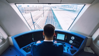 Person in Rückenansicht sitzt in Fahrerkabine eines Zuges: Bedient auf blauem Steuerungscockpit Tasten, vor ihr verschwommen Fenster mit Blick auf Gleise