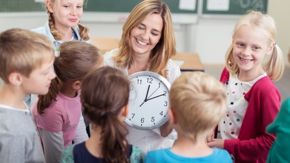 Person mittleren Alters hält eine Uhr in Händen und Kinder stehen ringsherum, im Hintergrund stehen Tische und eine grüne Tafel