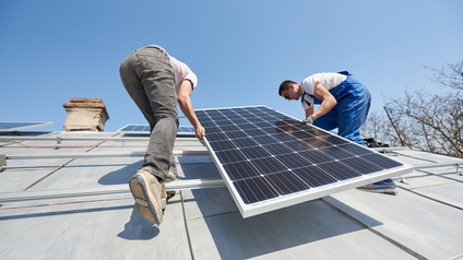 Zwei Personen bei der Montage einer Solaranlage am Dach eines Hauses unter blauem Himmel