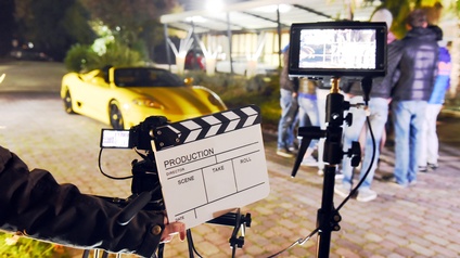 Detailansicht einer Hand, die schwarzweiße Filmklappe hält mit der Aufschrift Production, vor Kamera und Screen, im Hintergrund verschwommen gelbes Sportauto und Personengruppe