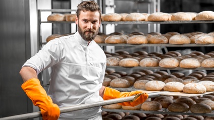 Person mit Bart mit gelben Schutzhandschuhen hievt Bäckerschaufel Brotlaibe aus Regal