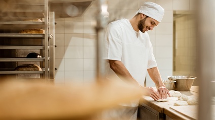 Lächelnde Person in weißem Gewand mit Mütze knetet Teig