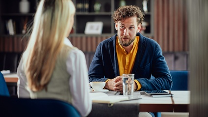 Zwei Personen sitzen sich an einem Tisch gegenüber. Die der Kamera zugewandte Person hat einen Bart und braune, kurze Haare. Auf dem Tisch sind ein Tablet und eine Tasse Kaffee. Im Hintergrund sind weitere Tische