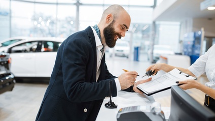 Lächelnde Person mit Bart unterzeichnet Unterlagen, andere Person reicht Autoschlüssel, im Hintergrund Autos