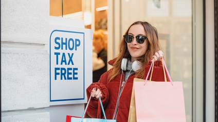 Eine Person mit Sonnenbrille, Kopfhörer und vielen Einkaufstaschen in beiden Händen steht vor dem Eingang eines Geschäfts. Neben ihr an der Wand ist ein Schild montiert, auf dem Shop Tax Free steht