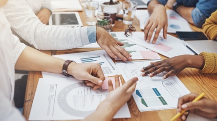 Um einen Tisch sitzen mehrere Personen. Auf dem Tischen sind viele Zettel mit verschiedenen Statistiken und Diagrammen. Die Personen deuten teilweise darauf