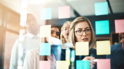 Fünf Personen stehen in einem Büro vor einer Glaswand. Auf der Wand kleben viele verschiedene, bunte Post-its. Eine Person hält einen Stift auf ein Post-it. Die anderen Personen blicken auf die Post-its