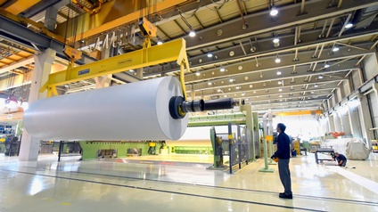 In einer sehr großen Halle ist eine sehr große, glänzende Papierrolle auf einer Metallstange, die über dem Boden steht. Daneben steht eine Person, die darauf blickt und eine Fernbedienung hält