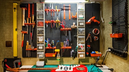 Eine rote, teilweise abgeblätterte E-Gitarre liegt auf einem Werktisch. Dahinter hängen an der Wand verschiedene Werkzeuge wie Schraubenzieher unterschiedlicher Art oder Zangen oder eine Bohrmaschine