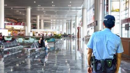 Rückenansicht einer Person in blauer Kleidung. Die Person steht in einer hohen Halle. In der Halle sind in der linken Hälfte Sitzreihen und eine Rolltreppen. Rechts sind lange Fensterfronten. Im Hintergrund sind Geschäfte