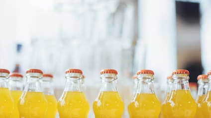Mehrere transparente Glasflaschen mit orangen Kronkorken und gelber Flüssigkeit nebeneinander stehend, Hintergrund verschwommen