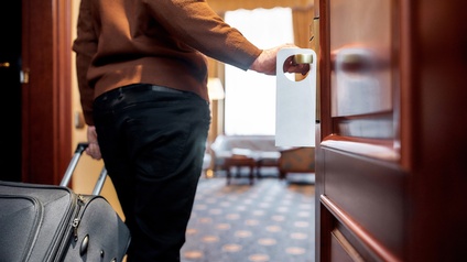 Eine Person, die mit der linken Hand einen Rollkoffer hinter sich herzieht, öffnet mit der rechten Hand eine Tür zu einem Hotelzimmer