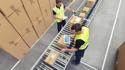 Aufnahme in Vogelperspektive: Zwei Personen in der Logistik scannen Pakete auf einem Fließband und führen Statistiken.