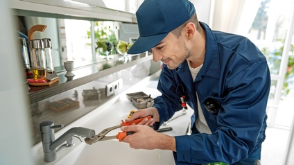 Eine Person in blauer Arbeitskleidung mit Kappe und Bart steht in einer Küche und hält eine Zange an einen Wasserhahn