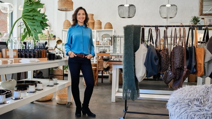 Unternehmerin mit blauer Bluse  steht lächelnd in ihrem Mode-/Geschenkegeschäft