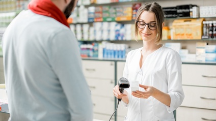 Frontale Aufnahme einer Person mit Brille, Kittel und geschlossenen Haaren, die hinter einem Tresen steht und in der rechten Hand einen Scanner hält. Ihr gegenüber steht eine weitere Person. Hinter ihr ist ein Regal mit vielen verschiedenen Produkten