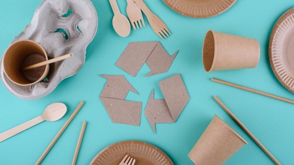 Mittig Recyclingzeichen − drei im abgerundeten Dreieck verlaufende Pfeile − umgeben von verschiedenen Verpackungsmaterialen wie Bechern, Tellern, Strohhalmen aus Papier und Einwegbesteck aus Holz auf türkisem Hintergrund