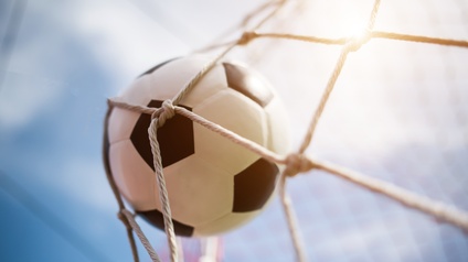 Detailansicht eines Fußballes - weißer Ball mit schwarzen Waben - der in ein weißes Netz fliegt, Hintergrund verschwommen himmelblau