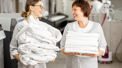 Zwei einander anlächelnde Personen in weißer Arbeitskleidung, eine Person hält Stapel weißer Morgenmäntel in Händen, die anderer Person trägt Stapel weißer Handtücher