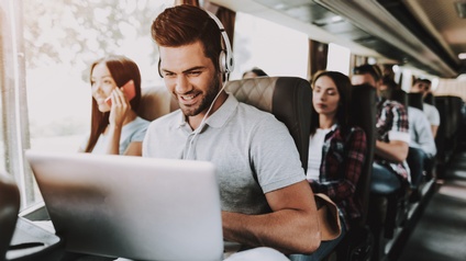 Lächelnde Person mit Kopfhörern sitzt in Bus und blickt auf Laptop, ringsum weitere Personen sitzend