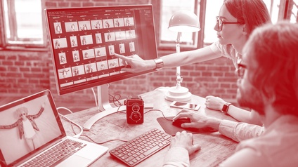 Zwei Personen blicken auf einem Monitor mit Bildern, der auf einem Schreibtisch steht, daneben steht ein aufgeklappter Laptop sowie eine leuchtende Bürolampe und eine alte Rollei Kamera