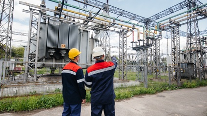 Zwei Personen in Arbeitskleidung und Schutzhelmen stehen vor einem umzäunten Umspannwerk. Eine Person hebt den Arm und zeigt auf Bauteile der Anlage. Im Hintergrund sind Transformatoren, Leitungen, Isolatoren und Metallkonstruktionen zu sehen