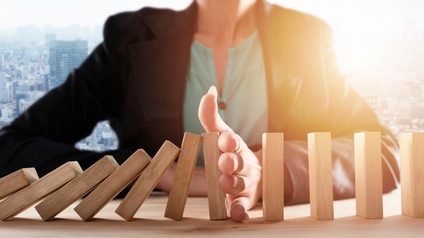 Die Büste einer Person ist zu sehen, sie sitzt an einem Holztisch. Mit der Hand stößt sie rechteckige Bausteine um. Im Hintergrund sieht man Umrisse einer Großstadt mit Hochhäusern. 