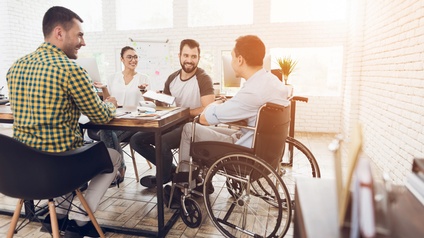 Um einem großen Tisch in einem Raum mit Glaswänden an einer Seite sitzen vier Personen. Eine Person sitzt in einem Rollstuhl. Die Personen reden miteinander. Hinter ihnen steht eine Flipchart 