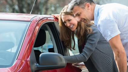 Zwei lächelnde Personen lehnen sich an rotes Auto und blicken zum geöffneten Fahrerfenster herein