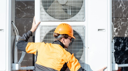 Rückenansicht einer Person mit schwarz-oranger Jacke, orangem Helm und Sonnenbrille, die neben einem Außengerät einer Klimaanlage steht und beide Hände auf dem Gerät hat