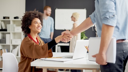Person sitzt in einem Raum an einem Tisch, lächelt und blickt zu einer anderen Person, die ihr die Hand gibt, im Hintergrund unterhalten sich zwei weitere Personen 