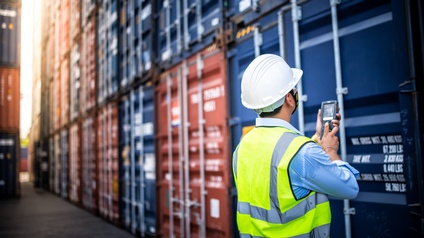 Rückenansicht einer Person mit Sicherheitsweste und weißem Helm, die mit den Händen einen Scanner hält. Direkt vor der Person steht ein blauer Container. Darüber und daneben sind weitere Container gestapelt.