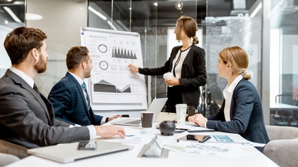 Personen in einem Besprechungsraum blicken gespannt auf eine analytische Chart, während auf ihrem Schreibtisch Laptops, Kaffeebecher sowie Statistiken liegen
