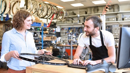 Eine Person steht an einem Tisch. Vor ihr sind ein Computer und ein Bankomatlesegerät. Links neben der Person steht eine weitere Person, die eine Fahrradspeiche hält. Im Hintergrund hängen von der Decke weitere Speichen.