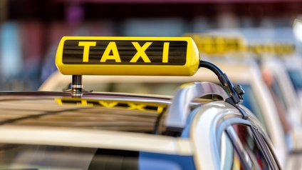 Leuchtend gelbes Zeichen auf Autodach: Gelber länglicher Leuchtkörper mit schwarzen Elementen und Schriftzug Taxi spiegelt sich in Autodach, im Hintergrund verschwommen weitere parkende Taxis