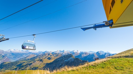 Sesselliftsessel mit Plexiglasverhüllung an Seil hängend unter blauem Himmel nähert sich gelber Station, unter Sessellift grüne Bergwiese, im Hintergrund Berglandschaft