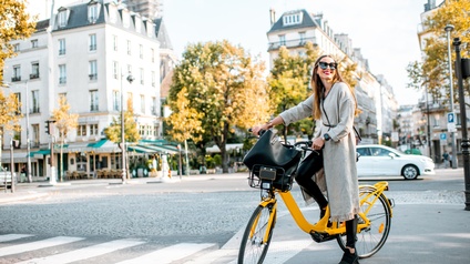 Lächelnde Person mit Sonnenbrillen posiert auf gelbem Fahrrad mit Korb, ringsum Stadtgeschehen