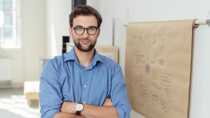 Selbstbewusste Person mit Brille und verschränkten Armen steht vor Whiteboard in einem Büro