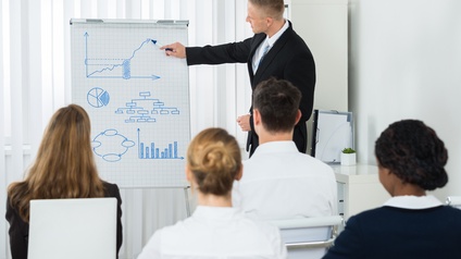 Person malt mit Stift auf Flipchart Diagramme, im Vordergrund mehrere Personen sitzend in Rückenansicht