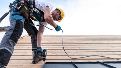 Eine Person mit Helm, Handschuhen und breitem Gürtel um die Hüfte steht auf einer schrägen Fläche aus horizontalen Holzbrettern. Die Person hält einen Bohrer auf eine Stelle. An ihrem Gürtel sind Karabiner befestigt