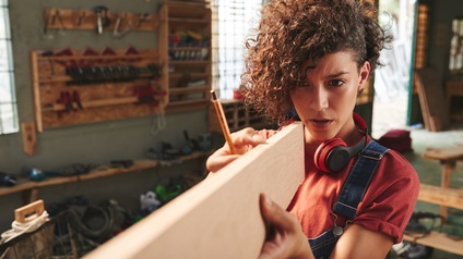 Eine Person mit Locken und Gehörschutz um den Hals steht in einer Werkstatt und hält ein Holzbrett nahe zum Gesicht und blickt auf die Kante. In der rechten Hand hält sie einen Bleistift. Im Hintergrund ist verschiedenes Werkzeug
