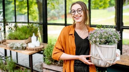 Porträt einer lächelnden Person mit Brille, die in einem Gewächshaus steht und einen Topf mit Lavendel hält. Im Hintergrund sind weitere verschiedene Pflanzen in Töpfen