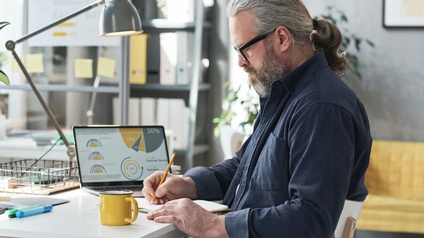 Person mit Bart und Brille sitzt an Schreibtisch und macht Notizen